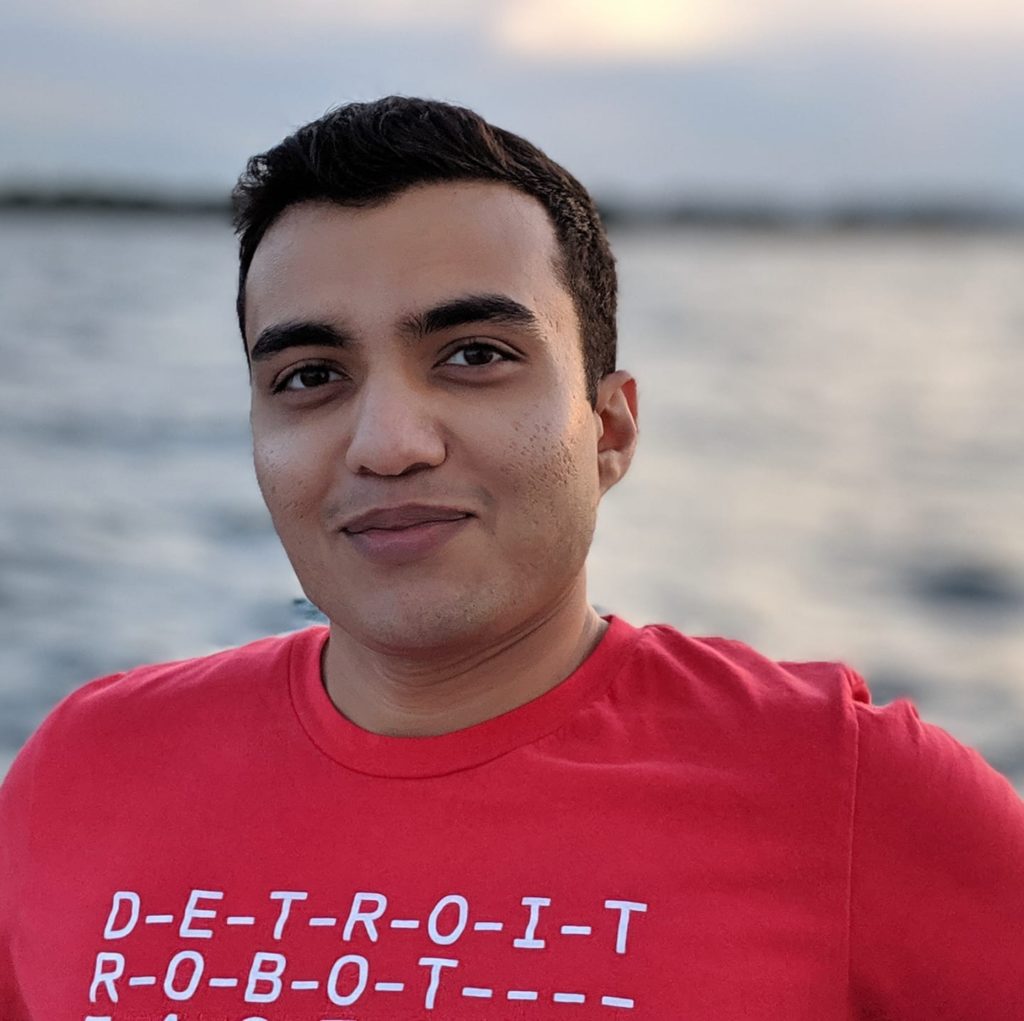 In this picture, Ninad is smiling warmly with the Detroit River in the background. The lighting is great and he is wearing a red Detroit Robot Factory t-shirt.
